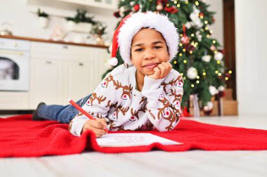 Noel Baba şapkalı bir çocuk rüya gibi gülümsüyor ve Noel Baba 'ya beyaz bir çarşafa bir mektup yazıyor. Bir Noel ağacının arka planında bir dilek listesi..