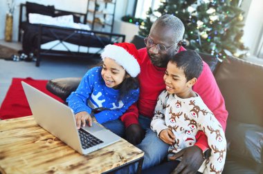 Afrika kökenli Amerikan ailesi - Noel Baba, oğul ve kız Noel Baba kazakları ve Noel Baba şapkalı kucaklaşma, gülümseme ve Noel ağacının arka planındaki akrabalarla video bağlantısı yoluyla iletişim.