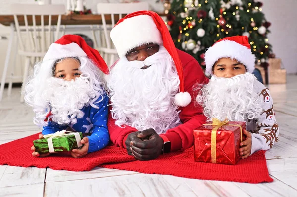 a family of African Americans - dad, daughter and son in Santa costumes and with gifts in their hands lie on a red plaid and smile against the backdrop of a Christmas tree and decor