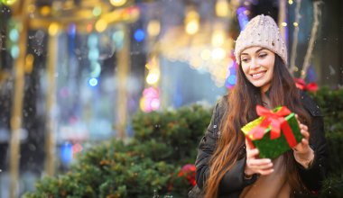 Ellerinde bir Noel hediyesi tutan ve gülümseyen örgü şapkalı genç bir kadının portresi. Arkaplanda kar ve Noel ışıkları var. Yazı, afiş, kopyalama alanı için alan.