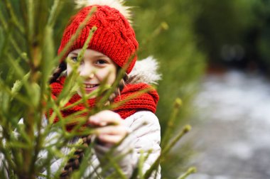 Kırmızı örgü şapkalı ve eşarplı küçük bir kız Noel ağacı pazarının arka planında gülümsüyor ve bir Noel ağacı seçiyor.