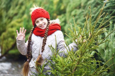 Kırmızı örgü şapkalı ve eşarplı küçük bir kız Noel ağacı pazarının arka planında gülümsüyor ve bir Noel ağacı seçiyor.