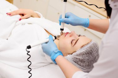 A cosmetologist conducts microcurrent facial therapy for a young woman using a device in a beauty salon. Hardware cosmetology, skin care, rejuvenation, regeneration.