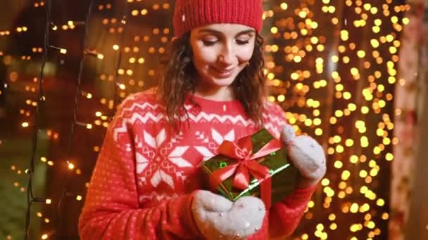 ニットの赤い帽子の若い可愛い女性と北欧のプリントのセーターは クリスマスライトを背景に彼女の手と笑顔で贈り物を保持します — ストック動画