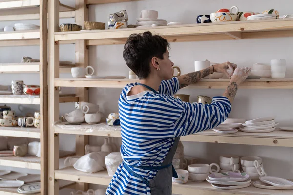 女性陶芸家が工房で仕事をしながら棚に手作りの陶器を置き 女性陶芸家が焼成前に棚に陶器や粘土のオブジェクトを乾燥させ アートスタジオの木製棚の近くに立つ — ストック写真
