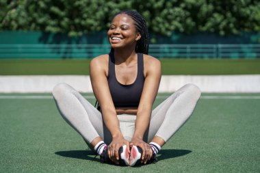 Mutlu Afrikalı Amerikalı kadın spor stadyumunun çimlerinde oturur ve bacak esnetme egzersizleri yapar ve ince gövdeyi destekler. Spor kıyafetleriyle gülümseyen siyah kız antrenmandan önce ısınma turlarında poz veriyor.