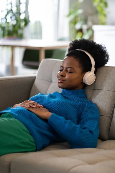 stock image Resting young African American woman in headphones with slight smile lies on sofa falling asleep to classical music. Relaxed lady sleeps after hard working day listening to favorite radio station