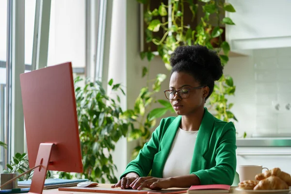 成功的非裔美国妇女担任编辑商业杂志打字的工作 坐在电脑桌前 戴眼镜的女孩带着植物在家里的办公室里为互联网出版社做远程工作 — 图库照片
