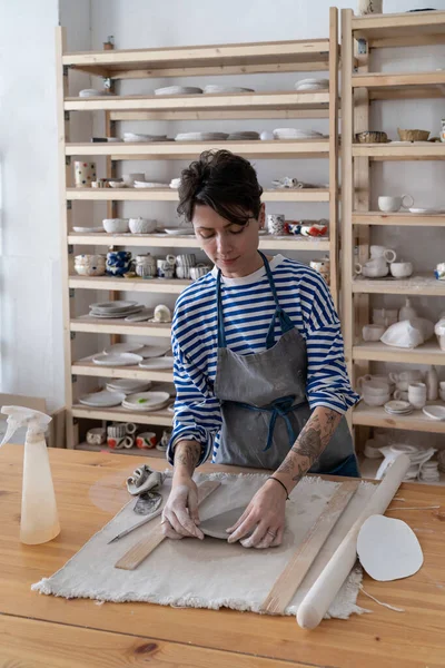 陶芸家の女性は粘土からのスタジオの彫刻プレートにテーブルに立っている ワークショップでアートを制作した後 ビジネスウーマンはリラックス 手工芸品店やショップビジネスのための手作り工芸品生産 — ストック写真