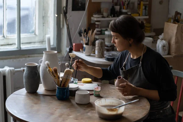 在车间工作的专心致志的妇女陶工坐在桌旁 用彩色油漆装饰陶器 做手工陶器的专业女雕塑家 风格为老式内饰 — 图库照片
