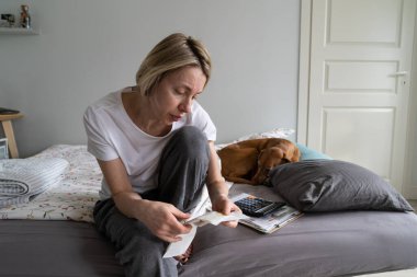 Eve geldikten sonra maliyet maliyetini doğrulamak için elinde süpermarketten gelen çeki tutan Avrupalı bir kadın. Ciddi bir kadın gündelik giysiler içinde yatağında uyuyan bir köpekle oturuyor. Gaz ya da elektrik faturaları okuyor.