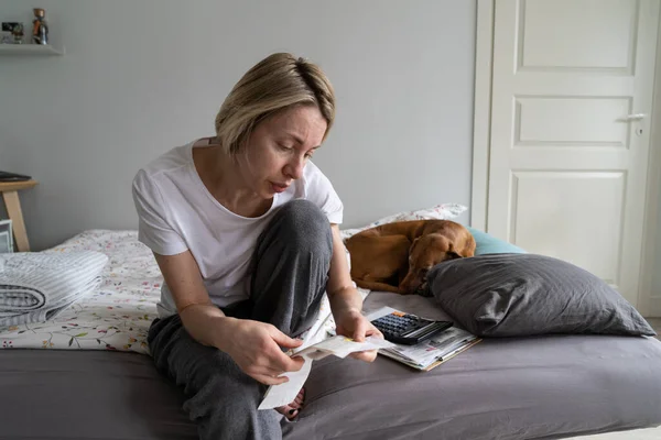 以欧洲女性为焦点 手握超市的支票 在回家后核对商品成本 身着休闲装的严肃女性和熟睡的狗一起坐在床上看书或电费 — 图库照片