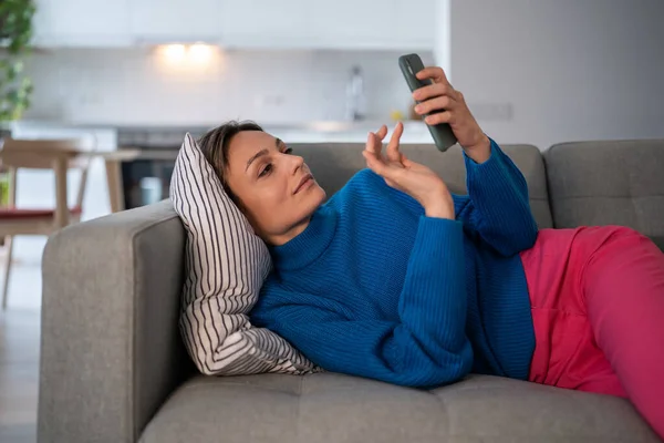 以轻松的年轻女性上网购物为重点 在下班后在舒适的沙发上使用手机应用程序聊天 拿着智能手机的女孩平静地享受着看屏幕的闲暇时光 — 图库照片
