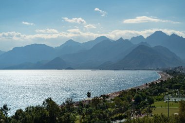 Turistler ve seyahat yolcuları için Antalya bölgesinde dağları ve deniz plajları olan resimli Türkiye. Gökyüzü manzaralı Türk tatil beldesi Konyaalti, sahil yakınlarındaki yüksek ağaçlar arasında otel binaları bulunuyor