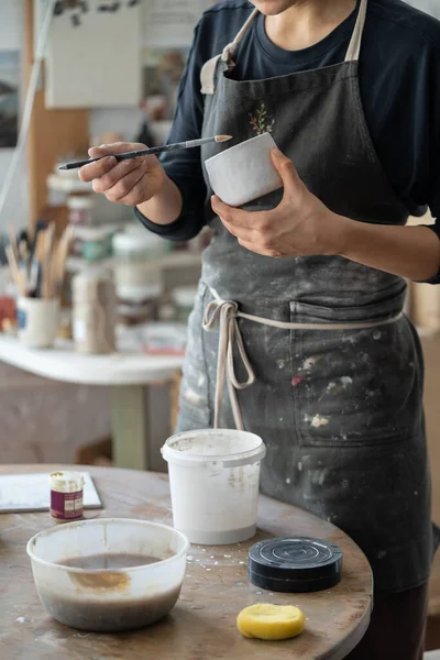 Creative craftswoman is engaged in manufacture of handmade ceramics standing near table in pottery studio. Cropped girl artist in apron holds brush and miniature vase makes dishes for souvenir shop
