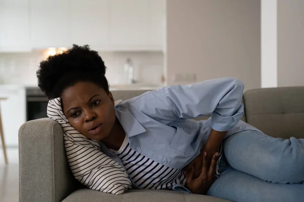 不幸なアフリカ系アメリカ人の女性が体調を崩し 鋭い痛みの後に手で胃をつかむ 若いです女の子は月経中や月経や困難な妊娠中にけいれんを経験ソファに横たわっています — ストック写真