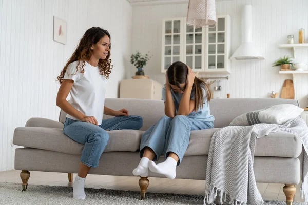 Strict woman looks at daughter sits on couch crying after ban on walks or home arrest. Little Caucasian girl was offended by mother after reprimand related to poor school performance or bad behavior.