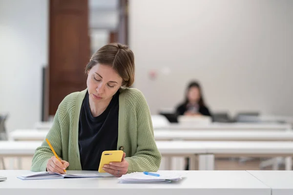 stock image Concentrated middle-aged scandinavian female student using smartphone for exam preparation while sitting at library table. Focused woman mature student studying, learning online, taking second degree