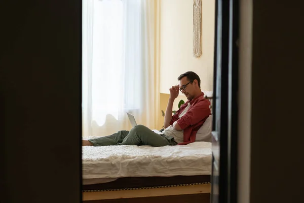 Задумчивый Запутавшийся Молодой Человек Фрилансер Очках Сидящий Кровати Глядя Экран — стоковое фото