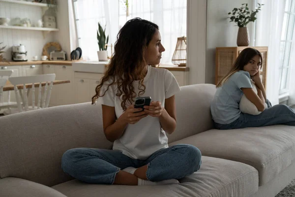 Caring puzzled mother sits on couch with mobile phone and looks back at offended daughter after punishment. Young reserved Caucasian woman in casual clothes wants to make call to child psychologist
