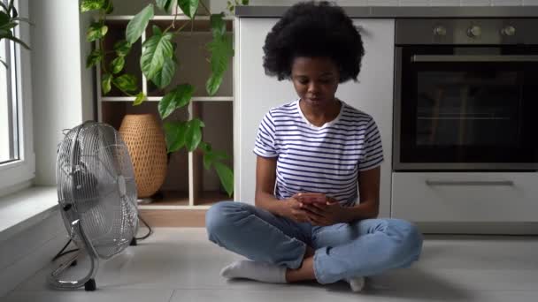 拿着电话的年轻卷曲的非洲裔美国女人坐在厨房的地板上 把电扇指向自己 穿着条纹T恤的女孩在手机应用程序上花时间与朋友聊天 — 图库视频影像