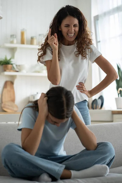 Angry mad woman mother disciplining-teen girl daughter sitting on sofa and covering ears not to listen. Woman talking to child with aggression, strict frustrated parent scolding teenage kid at home