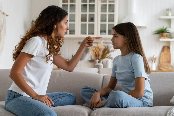Nervous Caucasian woman looks with anger at guilty daughter who does not want to obey parents or behaves badly. Family parenting process with single mother pointing finger at teen girl sits on sofa