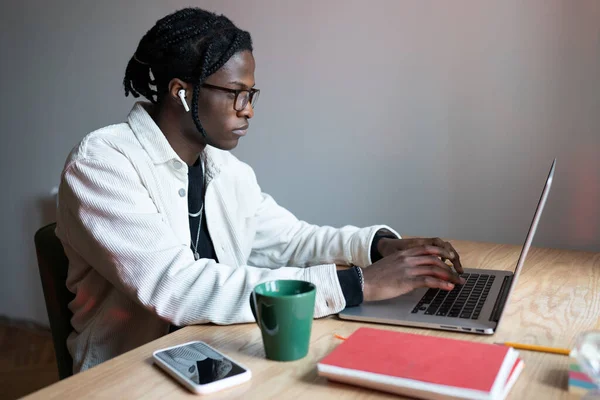 Carried away creative African American man in headphones sits at wooden table with laptop getting internet education. Young focused guy in stylish casual clothes doing extracurricular work for college