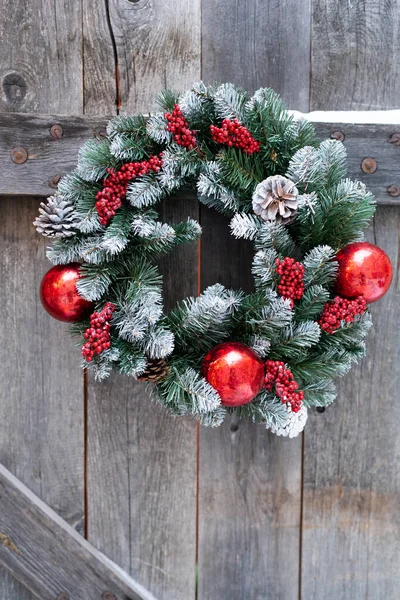 Christmas wreath of spruce twigs made of glass balls of red color hanging on wooden fence of village house. Close-up new years decorations for gate to country cottage to decorate for winter holidays