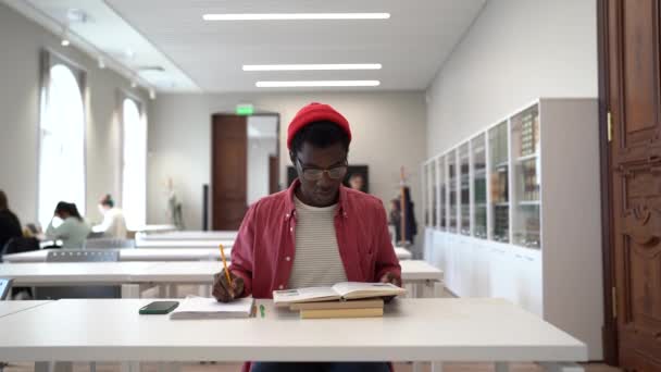 図書館で勉強するアフリカ系男性の学生に焦点を当て 要約を書くか 宿題をする一方で 彼の女性の友人の同級生は彼にコーヒーカップをもたらします 2人の多人種の学生と女性が一緒に勉強する — ストック動画