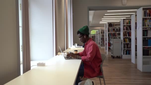 図書館でノートパソコンに取り組んでいる若いアフリカ系アメリカ人の男性は 勉強に疲れ ウェビナーを遠隔で見て勉強している黒人の学生を疲れさせ 研究作業に取り組んでいます オンライン学習疲労 — ストック動画
