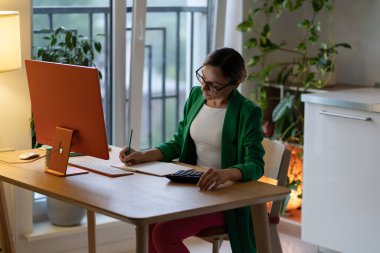 Gözlüklü düşünceli bir kadın not defterindeki iş bütçesi projesi için liste yapıyor. Bilgisayarlı Freelancer evden uzakta çalışmaktan hoşlanıyor. İş kadını muhasebe analizi faydaları yarattı