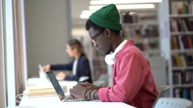 Odaklanmış Afrikalı hippi öğrenci gözlük takıyor çevrimiçi çalışıyor, dizüstü bilgisayarda yazıyor, araştırma çalışması hazırlıyor, kütüphanede masa başında oturuyor, seçici bir şekilde odaklanıyor. E-öğrenme, uzaktan eğitim kavramı