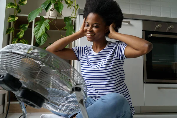 stock image Smiling African American woman using electric floor portable fan to maintain ideal indoor temperature during summer months, happy joyful young black female cooling herself enjoying fresh air at home