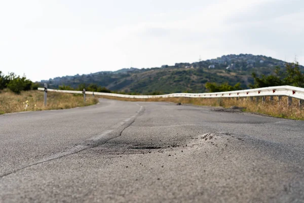 stock image Broken spot in middle of road stretching in wild area on sunny summer day. Deformed asphalt caused by heavy trucks on highway with traffic barrier built in countryside against high forestry mountains