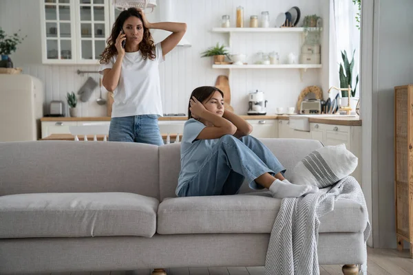 stock image Angry strict woman mother holding smartphone making phone call to husband to discuss teenage daughter behavior, upset teen girl sitting on sofa covering ears not to listen mom lecturing her at home