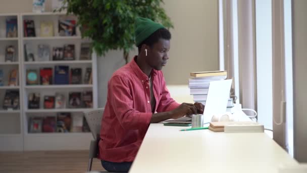 同僚のスペースでラップトップで働いている若いアフリカ人のフリーランサーは 電話に気を取られています 図書館でオンライン学習中に友人とオンラインでチャット黒の学生の男 学術的普及 — ストック動画