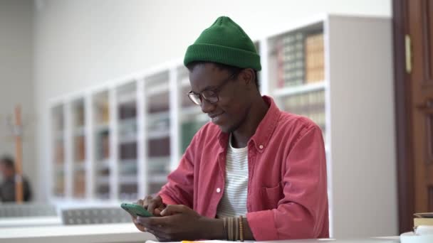 Улыбающийся Чёрный Парень Смартфона Сидящий Библиотеке Счастливый Молодой Африканский Студент — стоковое видео