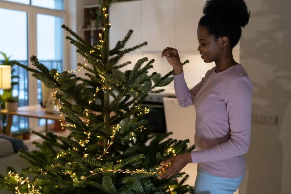 stock image Young happy smiling african woman decorating Christmas tree in minimalist way, cheerful black girl stringing LED garland along branches, preparing your home for holiday season. shallow depth of field