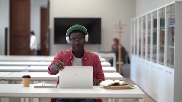 疲れたアフリカ人の学生はノートパソコンを閉じ ヘッドフォンを外し 図書館の机に座り 勉強から休憩を取り ストレッチの練習をした黒人青年を疲れさせた 燃え尽きた研究 — ストック動画