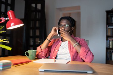 Stresli afro-amerikalı adamın gecikmiş iş süreleri hakkında telefon görüşmesi yapması. Şok olmuş serbest çalışan serbest çalışan serbest çalışan bir adam, masada dizüstü bilgisayarla oturuyor ve evinde kupa takıyor.