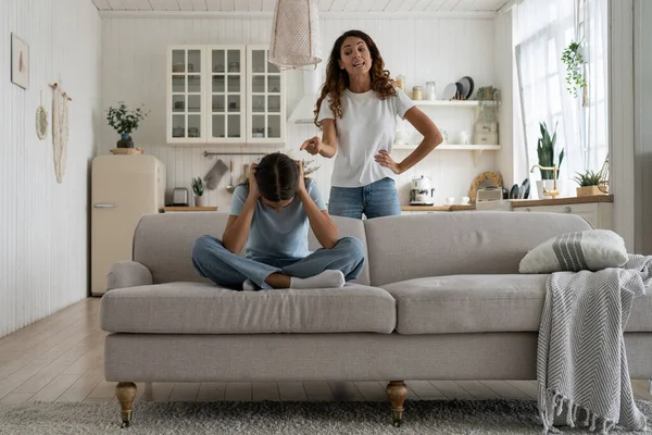 Strict mother scolding unhappy teenage daughter at home, mom lecturing teen girl, child sitting on sofa feeling afraid covering ears with hands. Parental harsh discipline, adolescent problem behavior