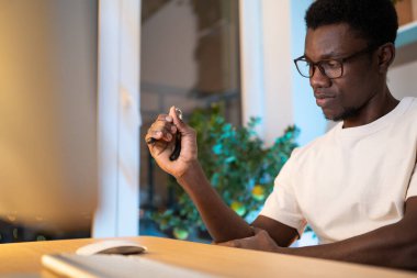 Young black worker in glasses trains hand muscles sitting at desk in living room at home. African American man in white t-shirt takes care of wrist using expander during long time work on computer