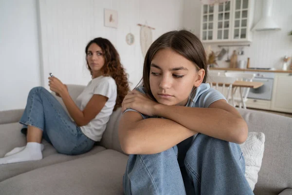Unhappy teen girl feeling sad after argument with parent, offended teenager sitting separately with mother on sofa at home, upset child having misunderstandings with mom. Teen depression concept