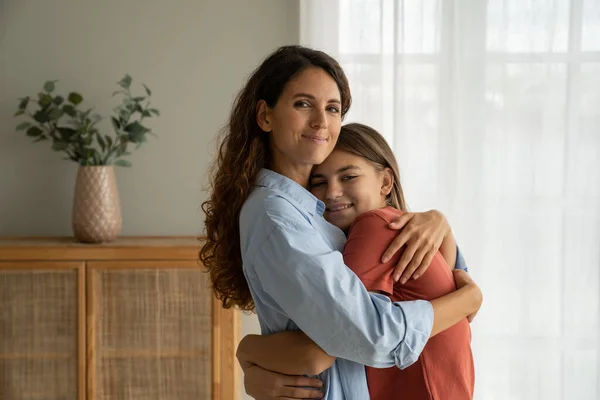 Kind optimistic Italian woman hugging teenage girl accepting congratulations on mother day. Little smiling daughter standing at window inside apartment cuddles up to mom when she misses very much