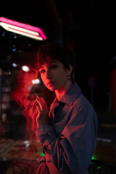 Sad lonely millennial woman with short hair in red neon light looking at camera, leaning on shop window at night, looking at camera, lonesome girl walking dark city streets alone. Neon photography