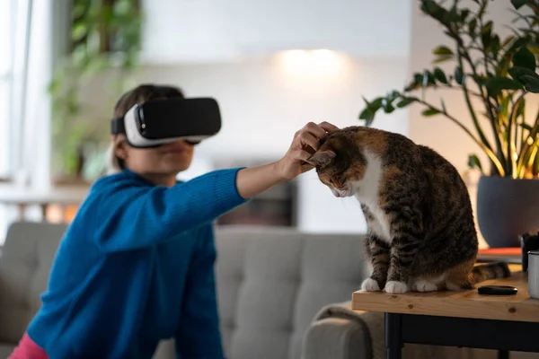 Positive woman in VR helmet petting cat sits on couch watching movie with 3D effects or virtual reality. Young Caucasian girl is fond of testing futuristic gadgets with AR technology function