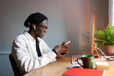 Kablosuz kulaklıklı genç Afrikalı öğrenci akıllı telefonu tutuyor, arkadaşlarıyla mesajlaşıyor, masaüstü bilgisayarıyla masaya oturuyor, evde ya da yurtta ders çalışırken telefonla gülümsüyor..