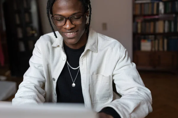 Smiling young African man in glasses using laptop at home, sitting at table chatting in social networks while spending leisure time. Happy black guy freelancer enjoying remote job, selective focus