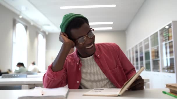 朗らかなアフリカ系アメリカ人男性の読書本を笑うことは 大学や高校の授業の合間にテーブルに座っています ケアフリー幸せな黒男楽しんで好きな趣味読書フィクションで図書館 — ストック動画